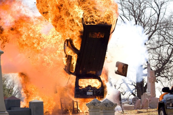 A dramatic explosion scene, featuring a car flipping mid-air in a cemetery, engulfed in flames.