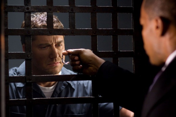 Nick Rice holds up a bracelet belonging to Clyde Shelton’s daughter in a tense prison scene. Gerard Butler, as Clyde, stares at the bracelet through the jail cell bars, symbolizing the emotional weight of his revenge-driven actions.