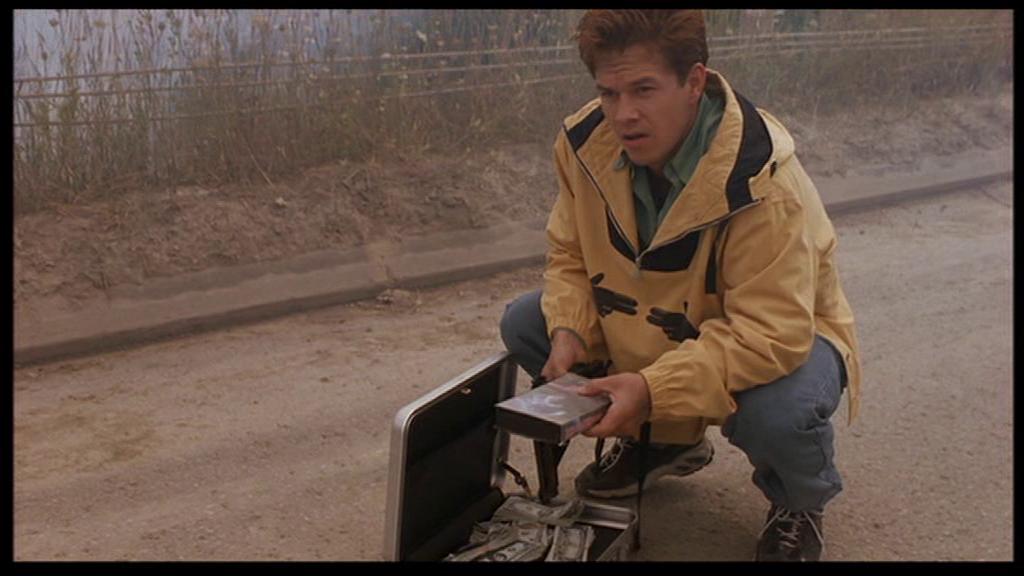 Mark Wahlberg as Melvin Smiley, crouched down outdoors in a yellow jacket, opening a briefcase filled with the ransom money.